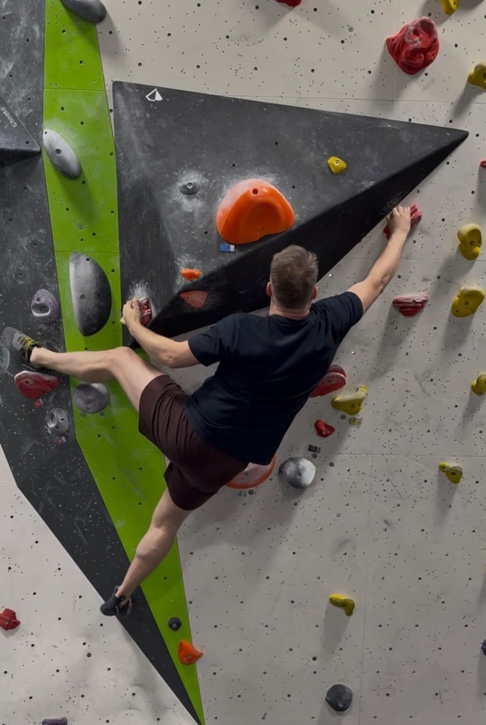Steven high on an indoor bouldering wall, spanning between two volumes.