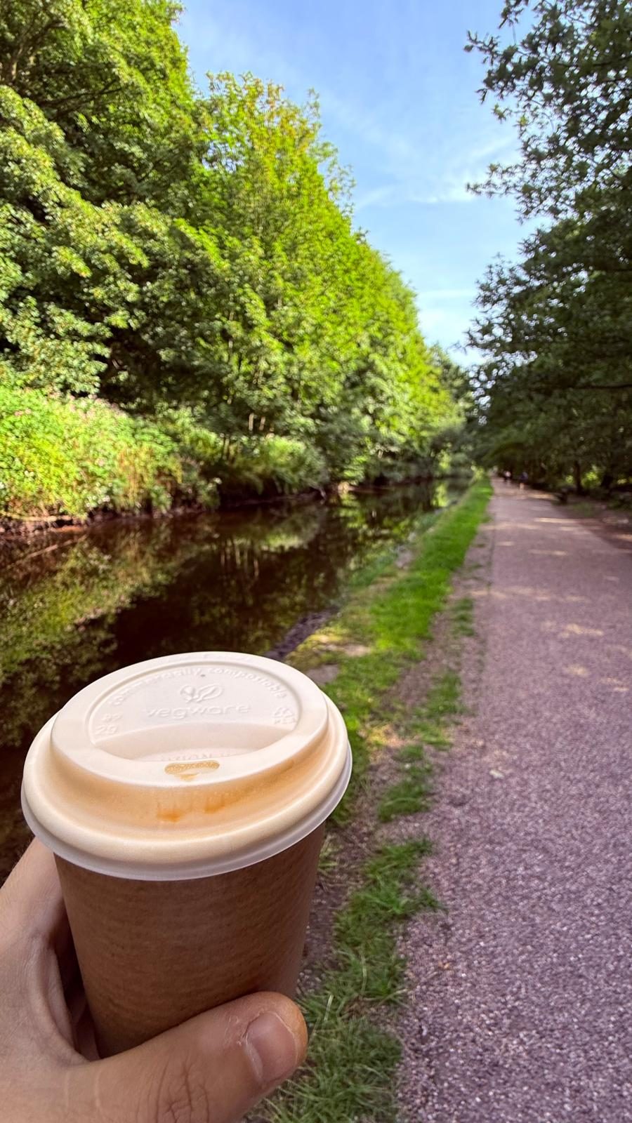 A canal towpath under trees, coffee cup held in shot.